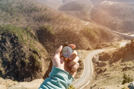 Close-up A mans hand holds a pocket compass against the backdrop of a mountain road and forest. The concept of outdoor navigationの写真素材