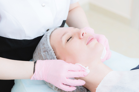 A close-up of the cleaning procedure in the office of cosmetology. The hands of the cosmetologist in pink gloves are removed from the face of a young girl with a sponge cleansing the maskの写真素材