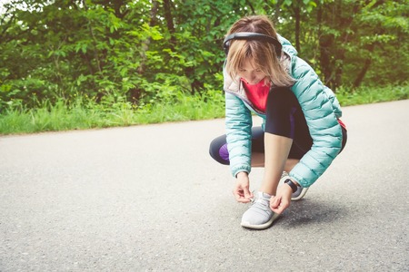 Portrait of a sporty blonde girl in headphones on a run in the forest. A girl sitting tied shoelaces on cross-country shoes. Active lifestyle. Sportの写真素材
