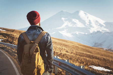 Bearded tourist hipster man in sunglasses with a backpack stand back on a roadside bump and watching the sunset against the background of a snow capped mountainの写真素材