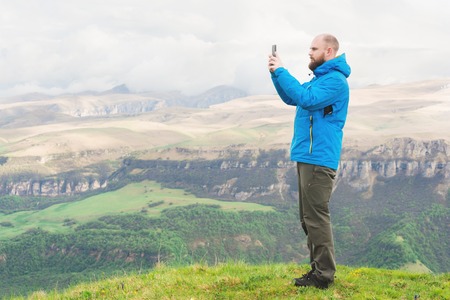 A bearded man in a membrane blue jacket stands in the nature against the backdrop of the mountains and takes pictures on the telephone landscapeの写真素材