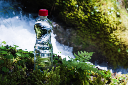 Transparent plastic A bottle of clean water with a red lid stands in the grass and moss on the background of a rugged mountain river. The concept of pure natural drinking waterの写真素材