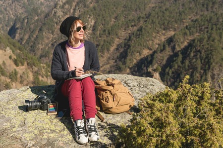 Stylish girl designer painter in the mountains sitting on a rock draws in a notepad at sunsetの写真素材