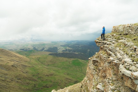 bearded hipster tourist man standing on the edge of a rock and looking out into the distance on an...
