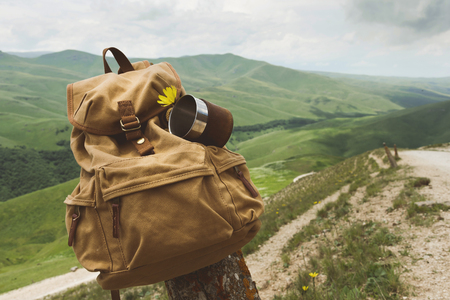 Hipster yellow vintage backpack with a mug fixed on it with a mug close-up front view. Travelers travel bag in the background of a mountain landscape and a roadの写真素材