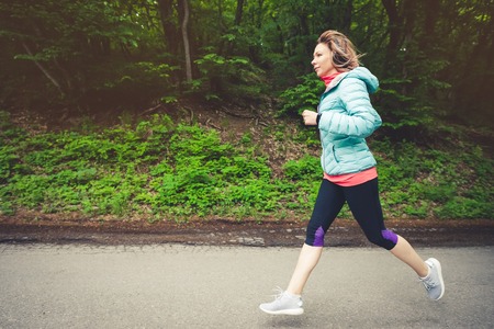 A young blonde woman running is practicing outdoors in a city mountain park in the forest. Warm rays through the branches of treesの写真素材