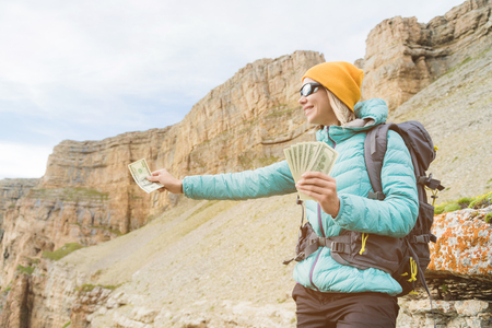 A traveler girl wearing a hat and sunglasses holds a hundred dollar bills in the hands of a fan against the backdrop of rocks on the nature. Handing money to nature and the windの写真素材