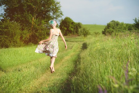 A cute cheerful young girl in a light cotton dress strolls along the country road against the backdrop of summer greenery at sunset. View from the backの写真素材