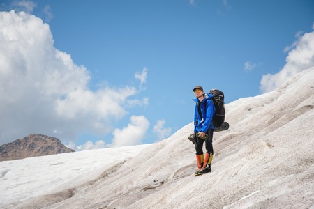 A mountaineer with a backpack walks in wheelchairs, stands on a dusty glacier with sneakers in the hands between cracks in the mountainsの写真素材