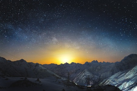 Moonset in the mountains at night with a horizontal milky way on the sky. Snow covered peaks of mountains at nightの写真素材