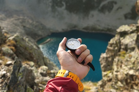 A mans hand holds a pocket magnetic compass for navigation against the backdrop of a rocky slope and...