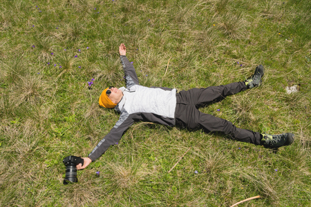 Happy girl photographer in sunglasses and with a camera in hand lies on the grass spreading arms and legs apart from the happiness of freedom on nature on the green grass in the mountainsの写真素材
