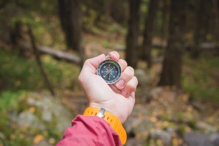 A beautiful male hand with a yellow watch strap holds a magnetic compass in the coniferous autumn forest. The concept of finding yourself the way and truthの写真素材