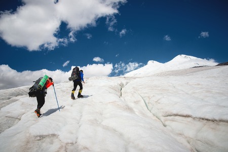 Two tourists, a man and a woman with backpacks and crampons on their feet walk along the glacier against the background of the mountain Elbrus and the sky and clouds. Back viewの写真素材