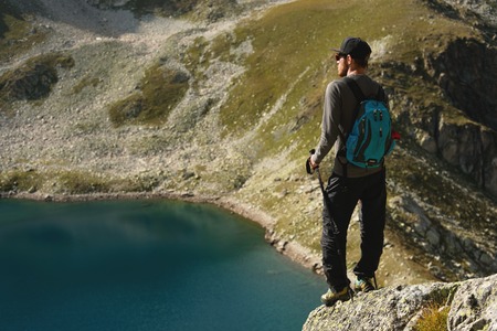 A bearded hipster tourist in sunglasses with a backpack is standing on the edge of a cliff high in...