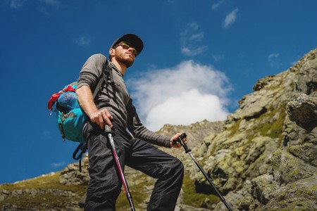 Porter of a stylish hipster traveler with a beard and a backpack in sunglasses and a cap with trekking poles standing on a rock against the background of a slope and blue sky in the mountainsの写真素材