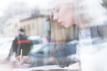 Portrait of an attractive young journalist girl with pen and notepad in cafe behind the showcase. Non-contrast view through window glass reflectionの写真素材