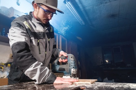 Young man with a beard in gray overalls and goggles by profession a carpenter works as an electric jigsaw in his home workshop. Cutting wooden partsの写真素材