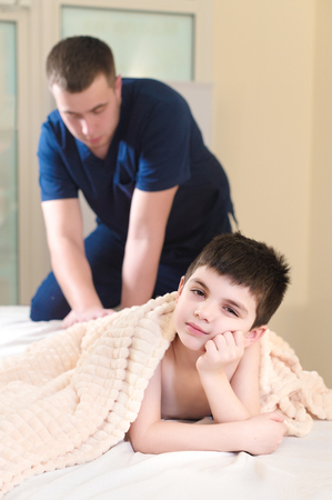 Male manual therapist massaging calves of a small patient. Little pensive boy at the reception of the procedure of massage in the office of physical therapyの写真素材