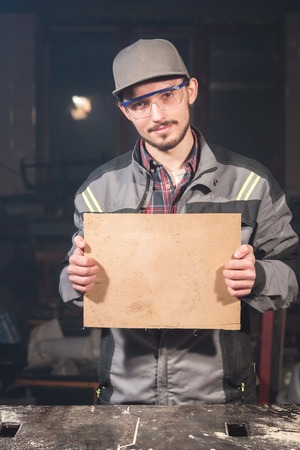 Portrait of a young carpenter in overalls and goggles with a mock up board holding a blank sign in his hands in his home workshopの写真素材