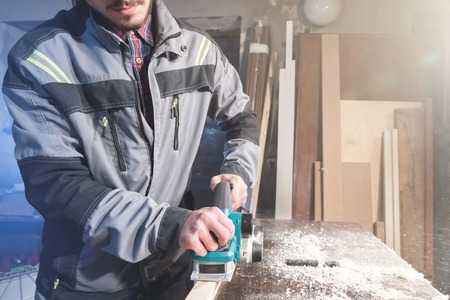 Young carpenter with a beard working with an electric plane with suction of sawdust. Leveling and...