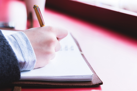 Close-up from behind the back of a young woman in a winter polto is holding a ballpoint pen and is going to write something down in a notebook on a red table. Notes.の写真素材
