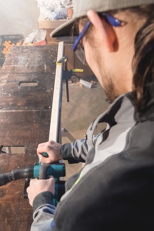 Portrait of a young carpenter working with an electrical plane in a home wood workshop. The concept of a business idea and a woodworking startup. Wide angleの写真素材