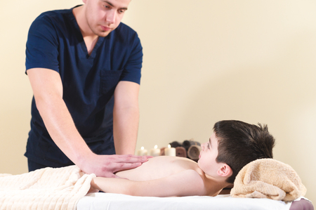 A male physiotherapist masseur makes a healing relaxing massage to a little smiling boy lying on a massage bed. Massage of the stomach and backの写真素材