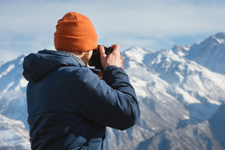 Portrait of a bearded male photographer in sunglasses and a warm jacket with a backpack on his back and a reflex camera in his hands takes pictures against the background of snow-capped mountainsの写真素材