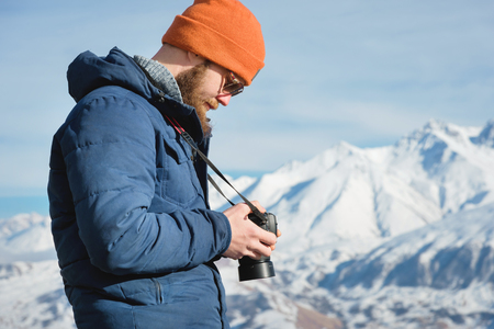 Close-up portrait bearded male photographer in sunglasses with dslr camera in his hands looks at the photos on the camera display on the background of snow-capped mountainsの写真素材