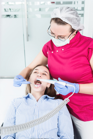 A professional dentist woman in glasses and overalls examines the oral cavity of a young girl in the dental chair using an intraoral stamotological video camera with LED illuminationの写真素材