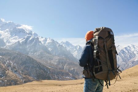 A bearded man in a hat and a backpack is standing outdoors against the snowy mountain peaks on a sunny day. Back viewの写真素材