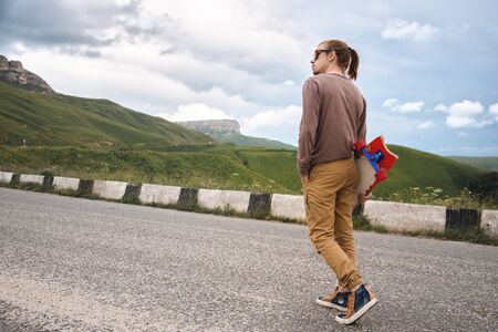 Young stylish hipster man with long hair and in sunglasses walking with a longboard in his hands on...