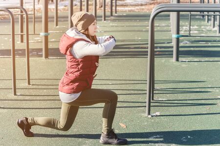 Portrait of a sports girl engaged in a workout in the winter on an outdoor sports field. Lunges and stitchesの写真素材