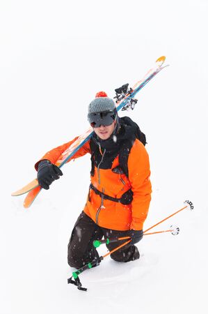Portrait of a professional skier athlete in a knitted hat and orange-black suit with a black ski mask with skis on his shoulder during a snowstorm on a light background in the snow. Wide angleの写真素材