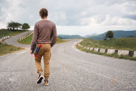 A stylish young man walks along a winding mountain road with a skate or longboard in his hands the evening after sunset. The concept of youth sports and travel hobbiesの写真素材