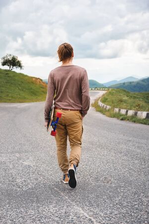 Young stylish man with long hair gathered in a ponytail and in sunglasses walking with a longboard in his hands on a country asphalt road. Mountains in the eveningの写真素材