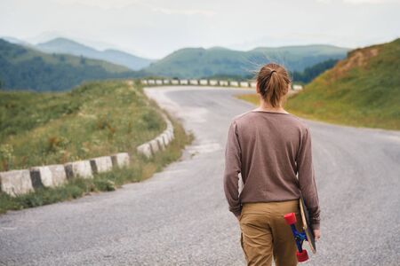 A stylish young man walks along a winding mountain road with a skate or longboard in his hands the evening after sunset. The concept of youth sports and travel hobbiesの写真素材