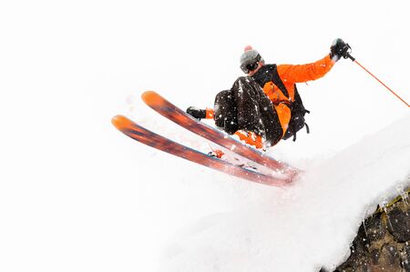 The athlete skier on a light background with a jump moves off the roof of a snow-covered hut with flying flakes of snow. The concept of winter ski sportsの写真素材
