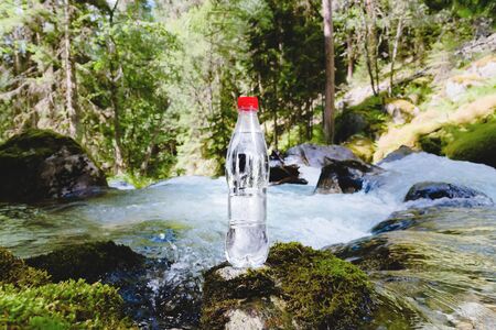 A plastic bottle with a red cap with fresh drinking water against a background of green forest and a mountain river stands on a stone with moss. The concept of pure natural mountain waterの写真素材
