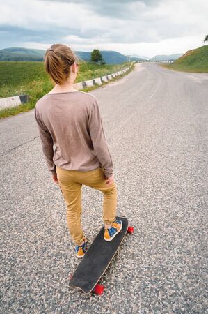 Young stylish man with long hair gathered in a ponytail and in sunglasses stands with a longboard on a country asphalt road in the mountains on the background of epic rocksの写真素材