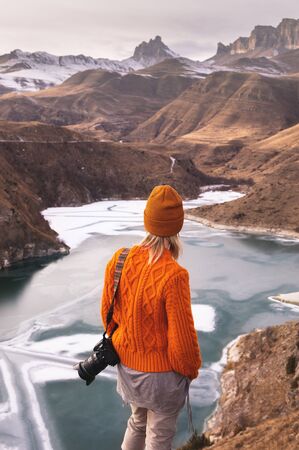 Portrait from the back of the girl traveler photographer in an orange sweater and hat with a camera...