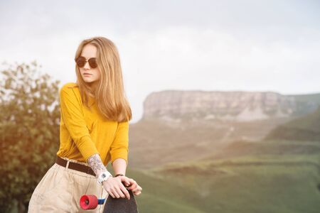 Beautiful and fashionable young woman in sunglasses and with a tattoo poses with a skateboard or...