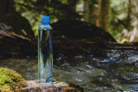 Plastic bottle blue cap with fresh drinking water on a background of green forest stands on a stone...