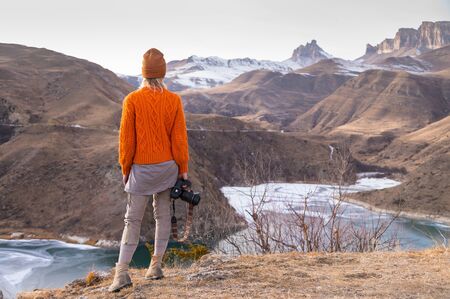 Portrait from the back of the girl traveler photographer in an orange sweater and hat with a camera...