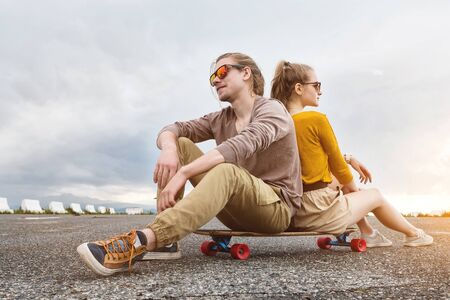 An attractive young couple in sunglasses sit back to back on their longboard in a suburban parking...
