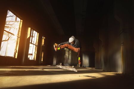 A young skater in a white hat and a black sweatshirt does a trick with a skate jump in an abandoned...