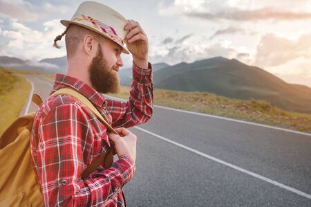 Portrait of a bearded happy serious traveler hipster with a backpack in a plaid shirt and a hat next to an unknown car stands on the road at sunset in the mountains. Happy and confident travel conceptの写真素材