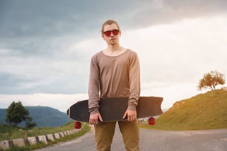 Stylish young man standing along a winding mountain road with a skate or longboard on his shoulder in the evening after sunset. The concept of youth sports and travel hobbiesの写真素材