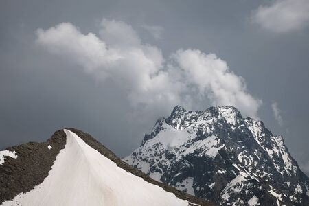 View of a high steep rocky mountain partially covered with snow against a dark cloudy sky with...